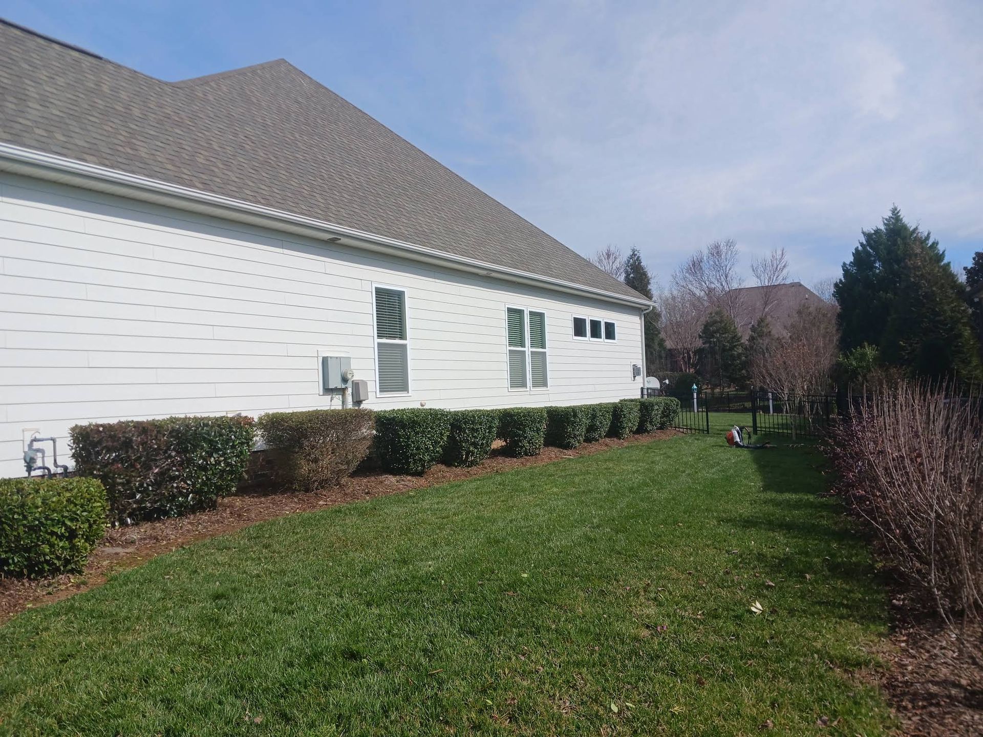 A white house with a gray roof and a side lawn bordered by a hedge under a clear blue sky.