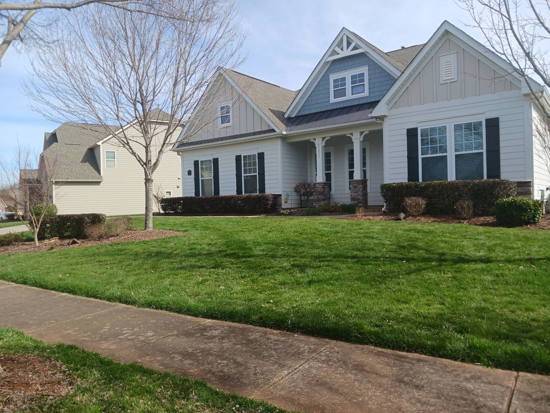 A suburban house with white siding, black shutters, and blue accents, featuring a manicured lawn and a concrete sidewalk.