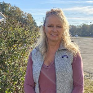 A woman wearing a patagonia vest and a pink shirt is standing in front of a bush.