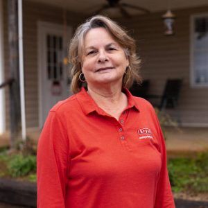 A woman in a red shirt is standing in front of a house.