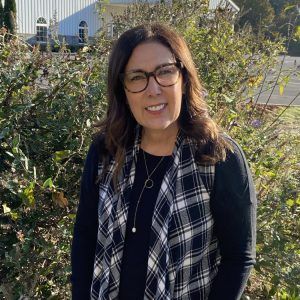 A woman wearing glasses and a plaid vest is standing in front of a bush.