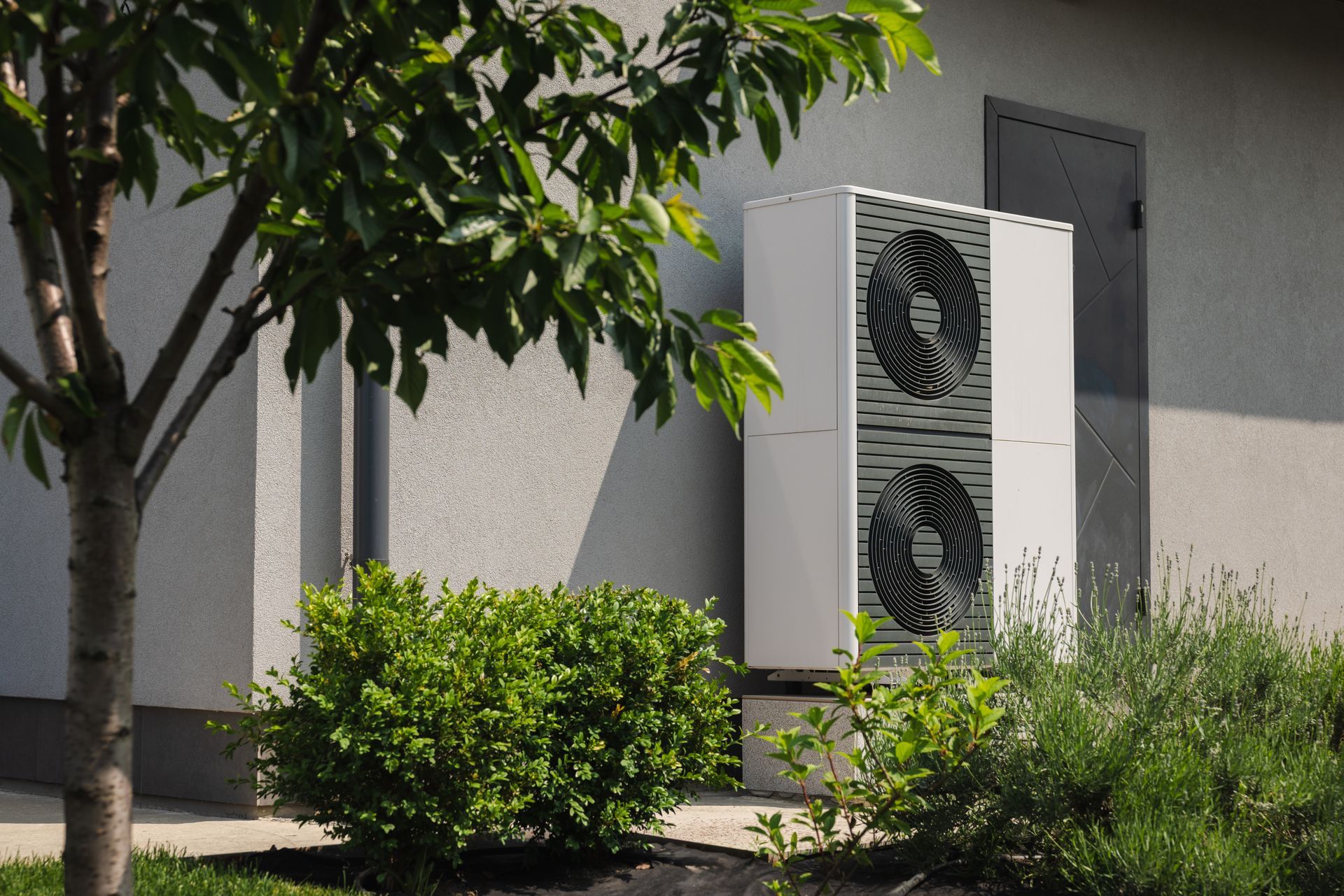 White heat pump unit outside a building with foliage and a tree.