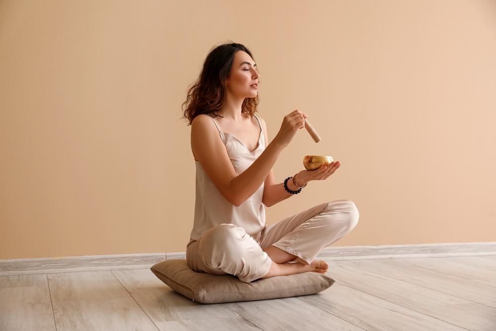 Woman seated cross-legged, using a singing bowl for meditation against a neutral backdrop.