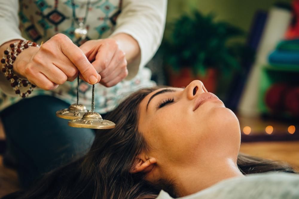 Woman receiving sound healing treatment with hand cymbals held over her head; eyes closed.