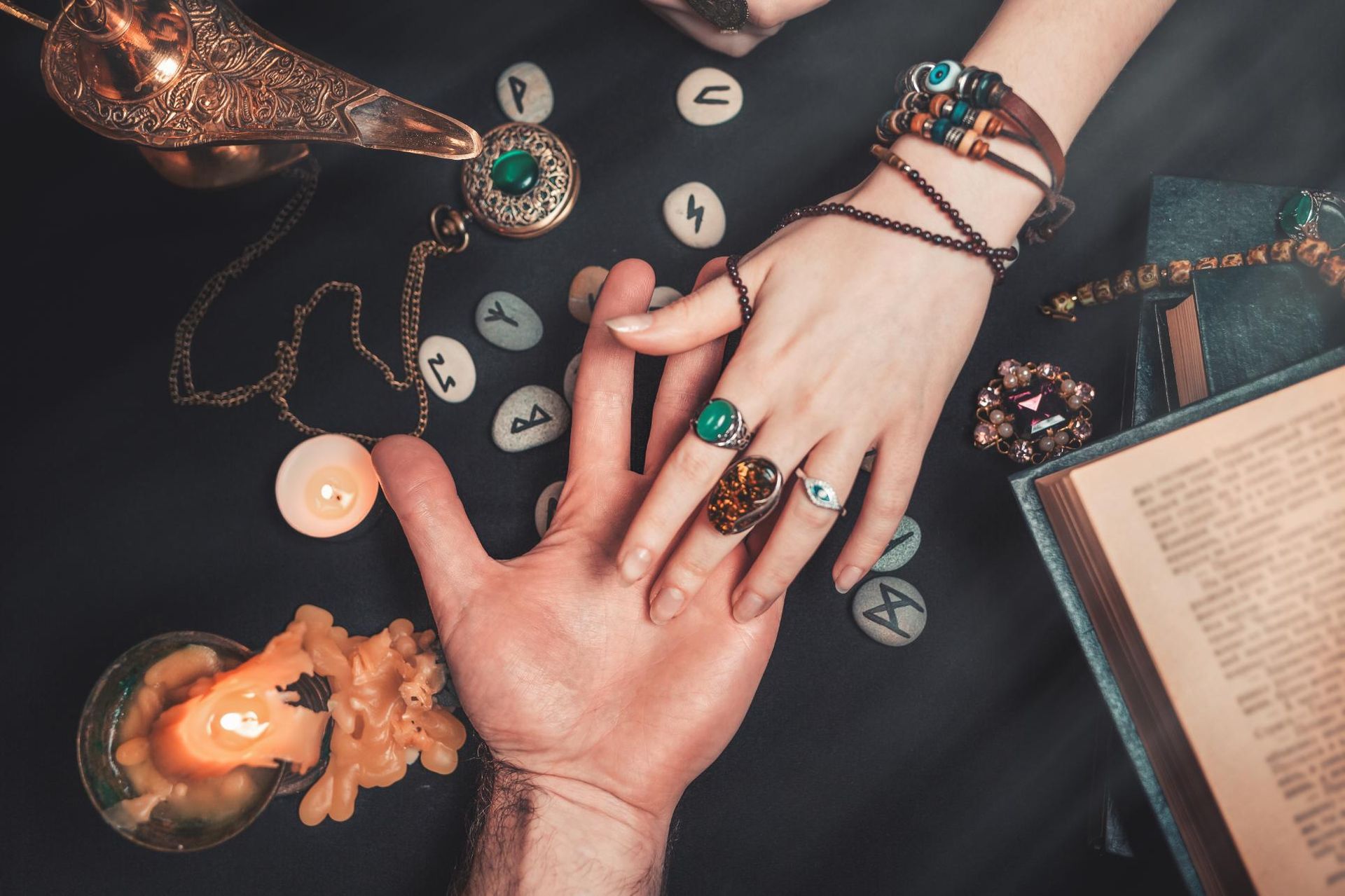 Hands in a palm reading session; candles, runes, and jewelry on a dark table.