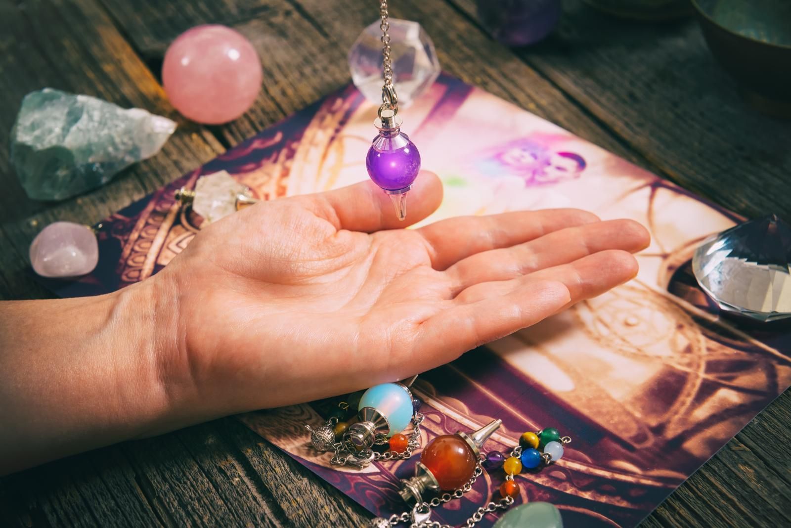 Hand holding a purple pendulum over a card, surrounded by crystals, on a wooden surface.
