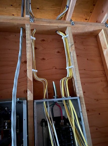 Electrical wiring cables running vertically along wooden wall studs into two metal electrical service panels.