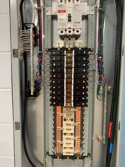 Open electrical breaker panel with a main circuit breaker, busbars, and rows of black switches connected by colored wires.