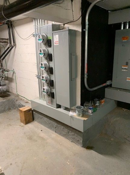 A row of five electric meters and an adjacent electrical panel mounted on a gray concrete base in a basement.
