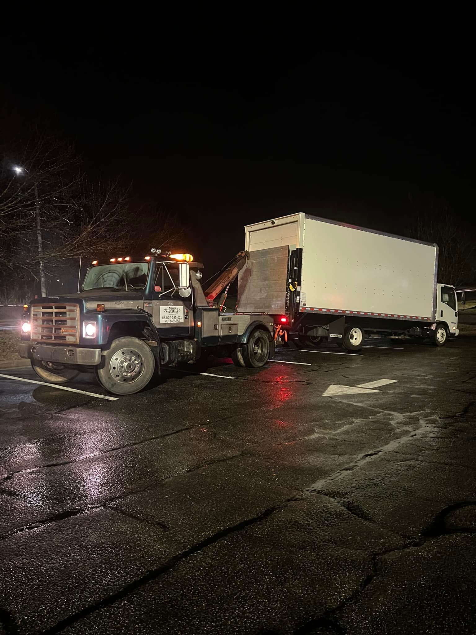 A tow truck is towing a white truck in a parking lot at night.