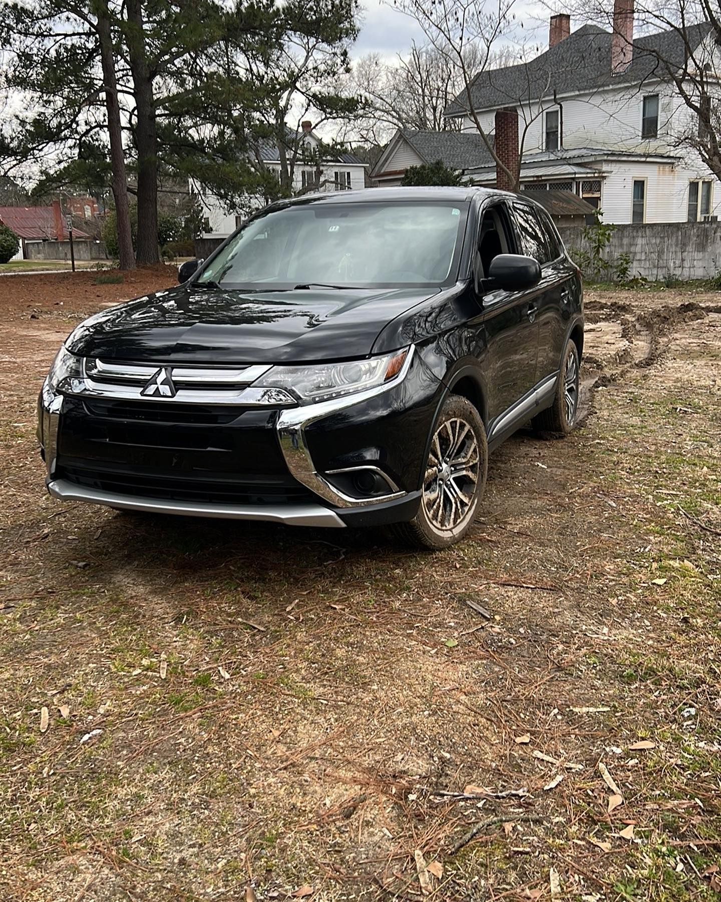 A black mitsubishi outlander is parked in a dirt lot in front of a house.