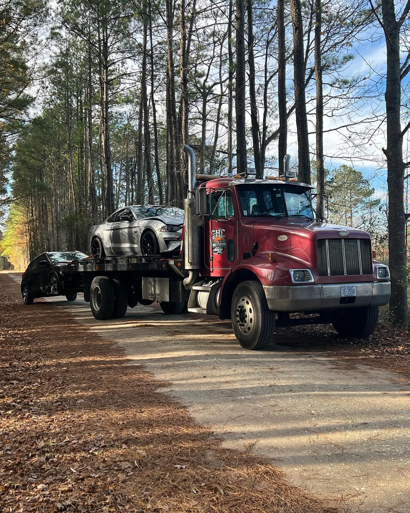 A red semi truck is towing a white car down a dirt road.