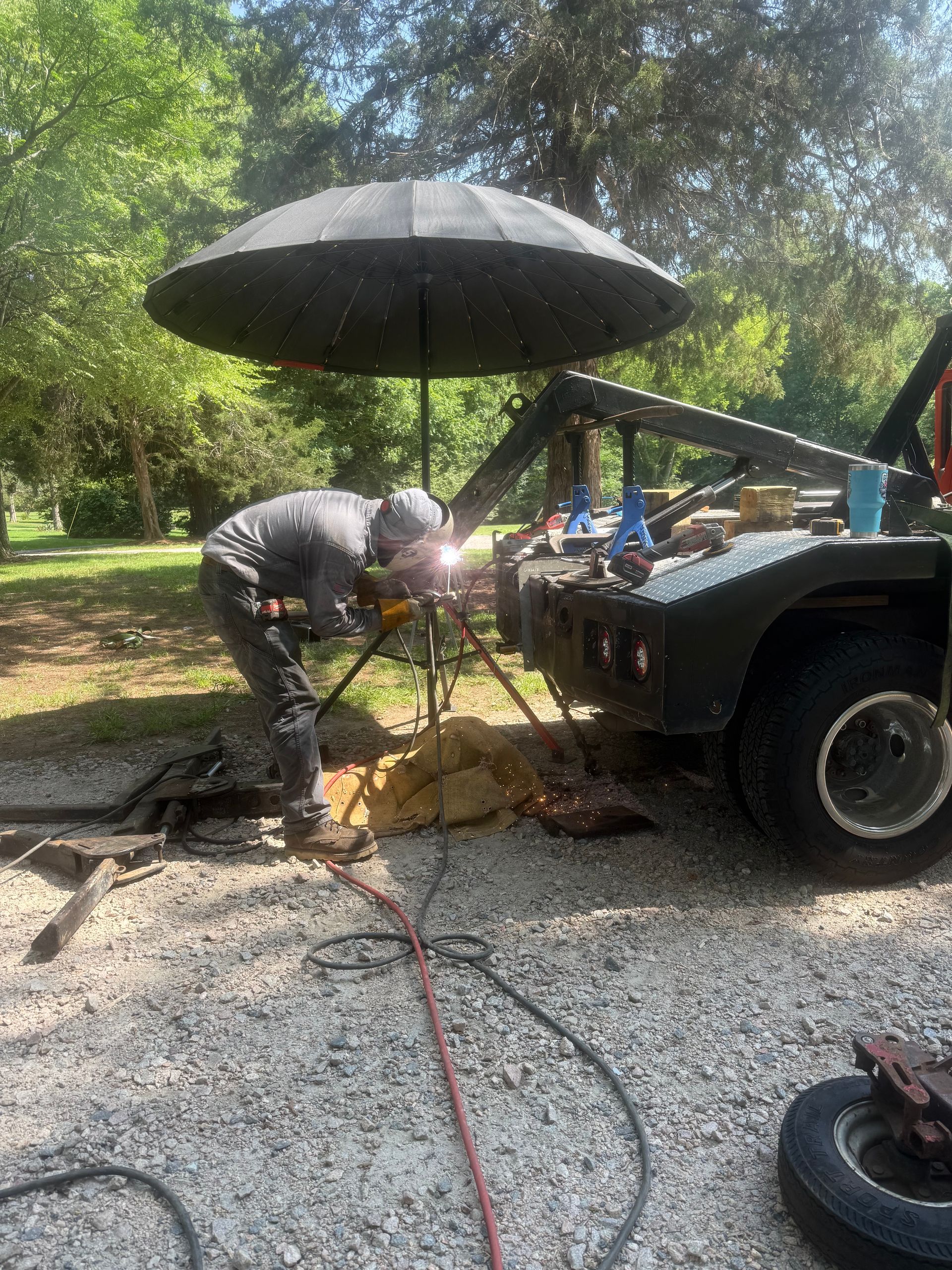 Person working under an umbrella beside a vehicle in a sunny gravel driveway.