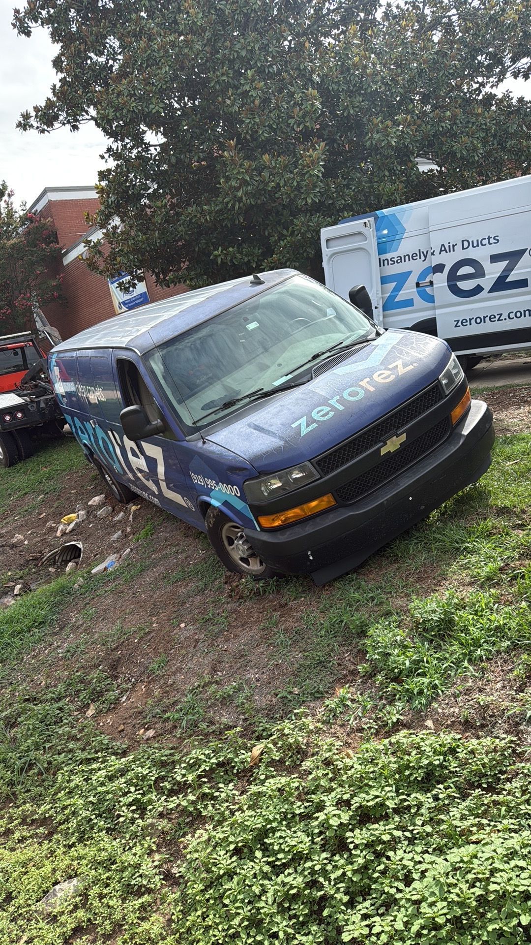 Blue ZEZ van parked on a grassy, muddy lot beside a white trailer and trees