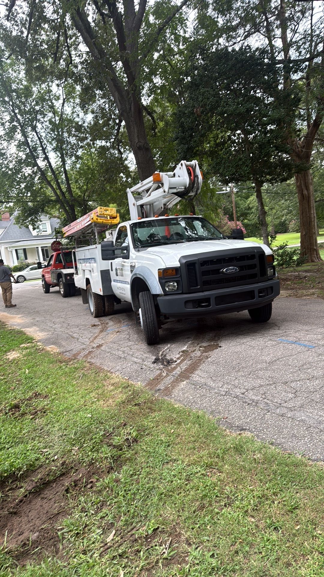 Two utility trucks on a tree-lined driveway, one with a bucket lift extended overhead.