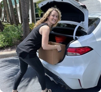 Woman smiling while loading a cardboard box into the trunk of a white car.