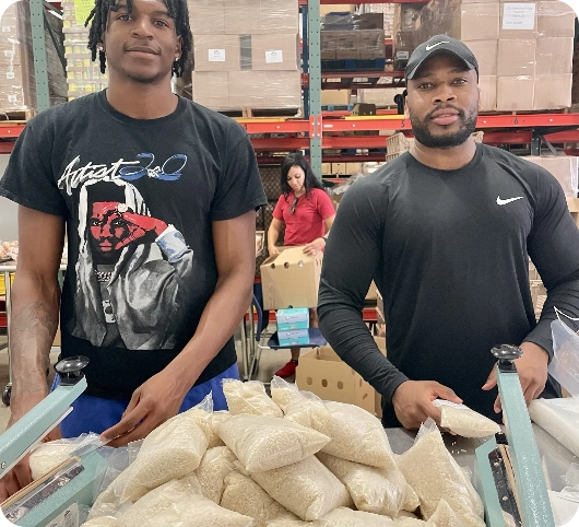 Two men packing bags of rice, with a woman in the background. Warehouse setting.