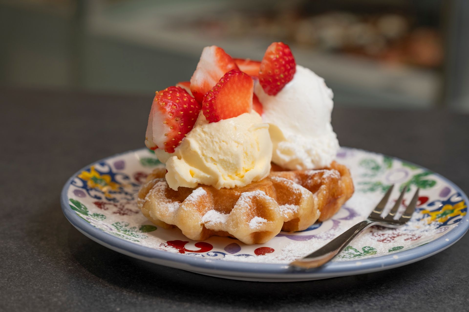 Waffle guarnito con panna montata, gelato e fragole, su un piatto floreale con forchetta.