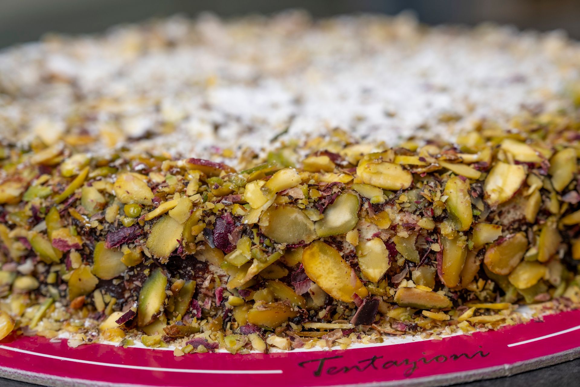 Primo piano di una torta rotonda ricoperta di pistacchi tritati e zucchero a velo su un piatto con bordi rosa.