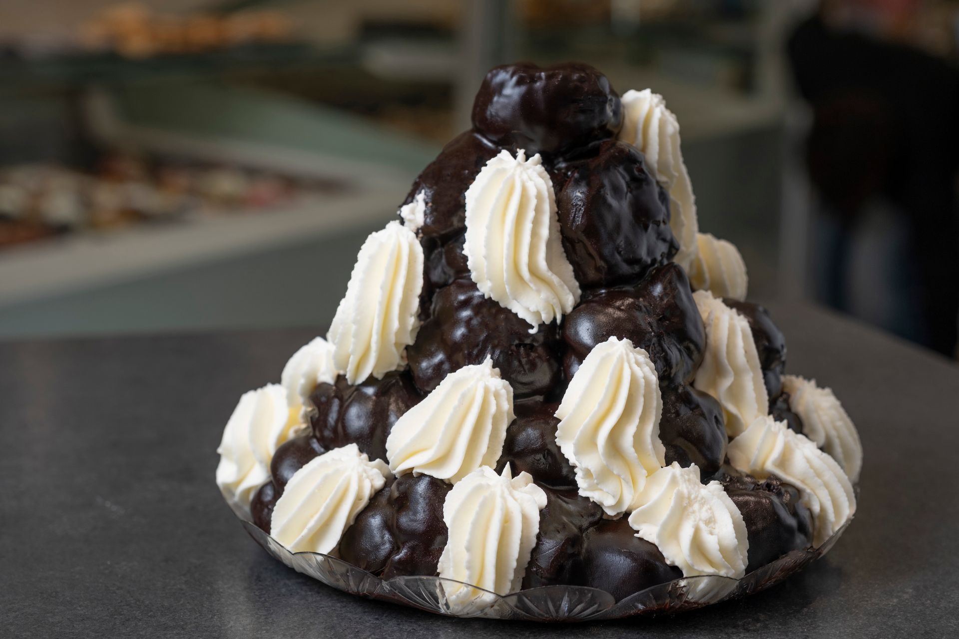 Torre di pasta croquembouche, profiteroles ricoperti di cioccolato fondente, con ciuffi di crema bianca, su una superficie grigia.