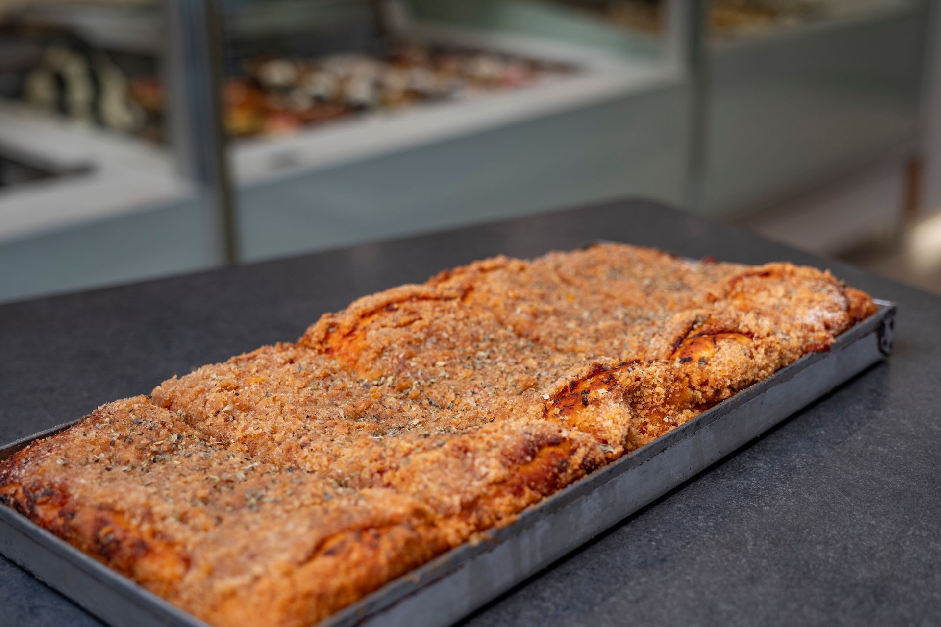 Una teglia rettangolare piena di prodotti da forno, dorati e ricoperti di zucchero, è appoggiata su una superficie scura in una panetteria.