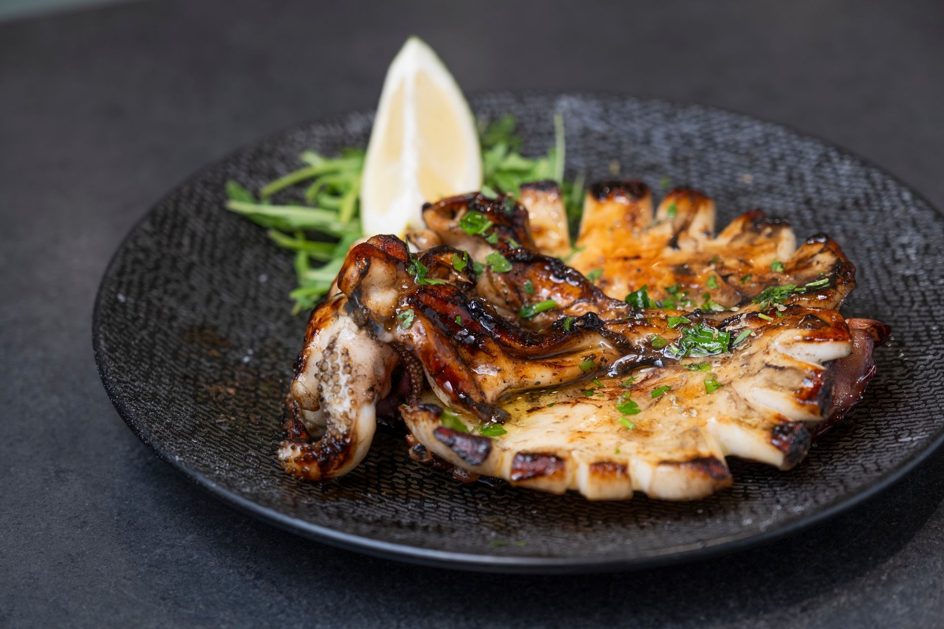 Calamari grigliati su un piatto scuro, guarniti con verdure e spicchio di limone.