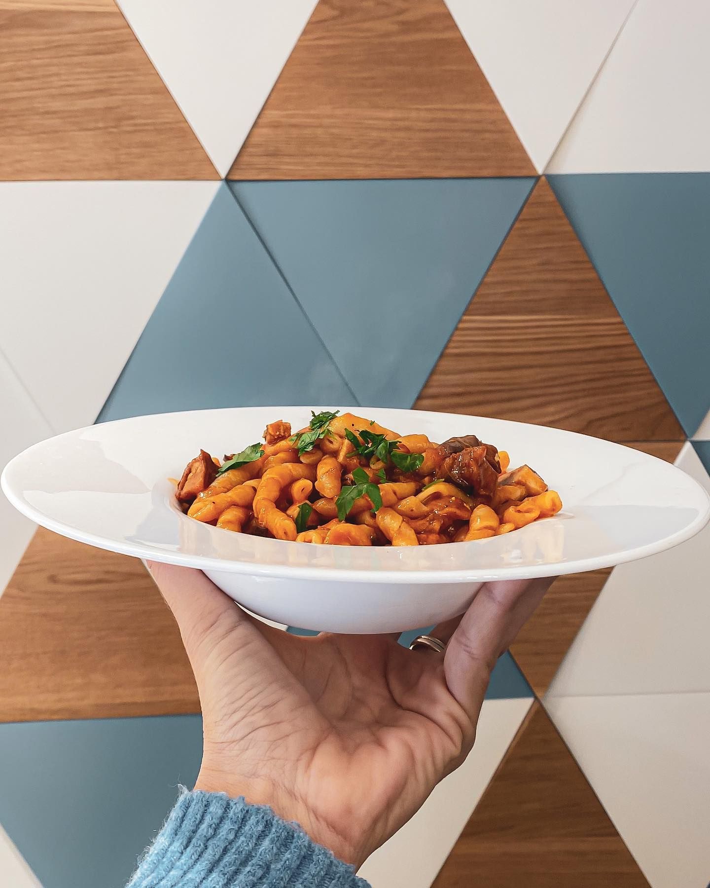 Mano che regge una ciotola bianca di pasta con salsa di pomodoro, chorizo e prezzemolo contro una parete geometrica.
