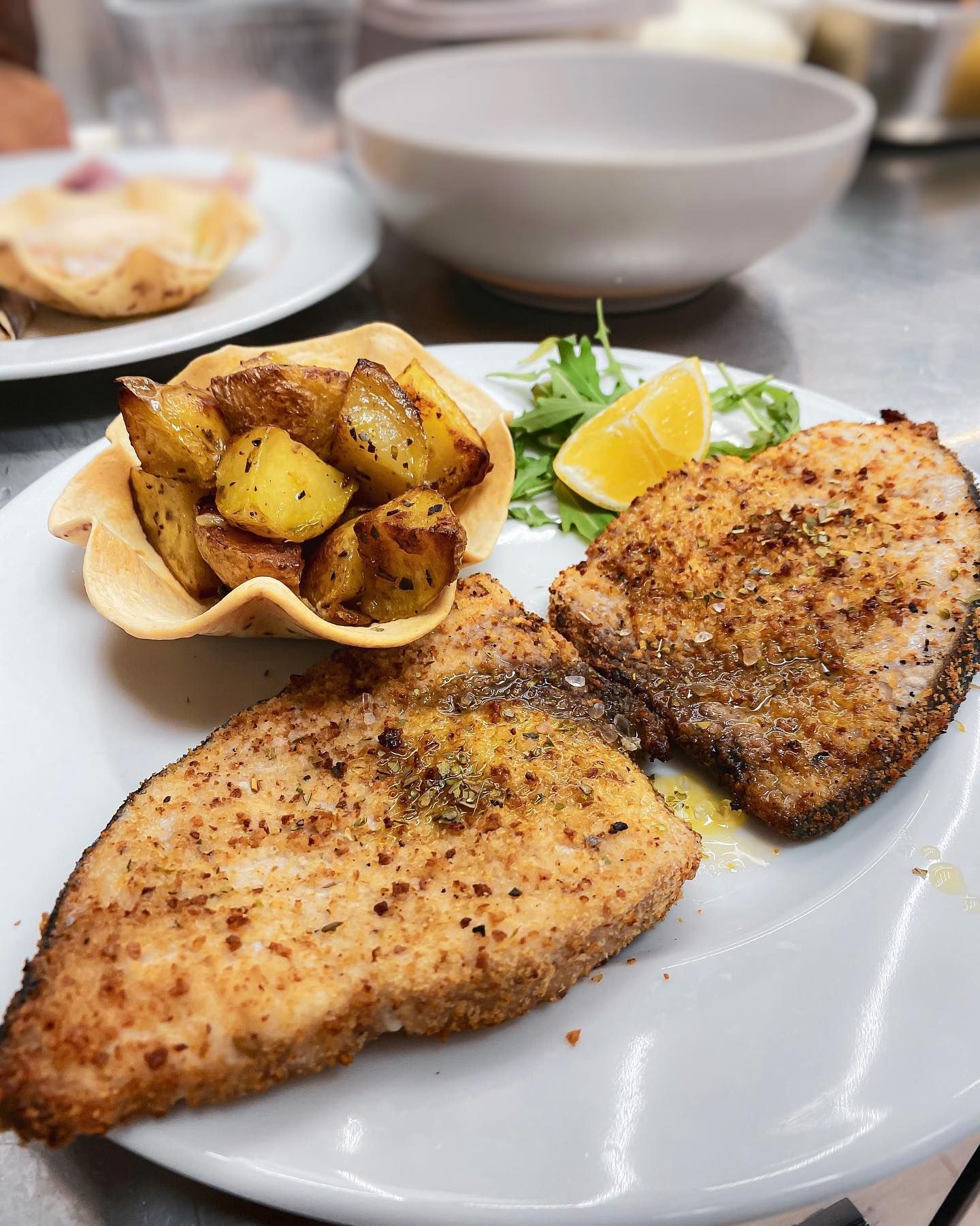 Pesce fritto in padella con patate arrosto in una ciotola intrecciata, spicchio di limone e verdure su un piatto bianco.