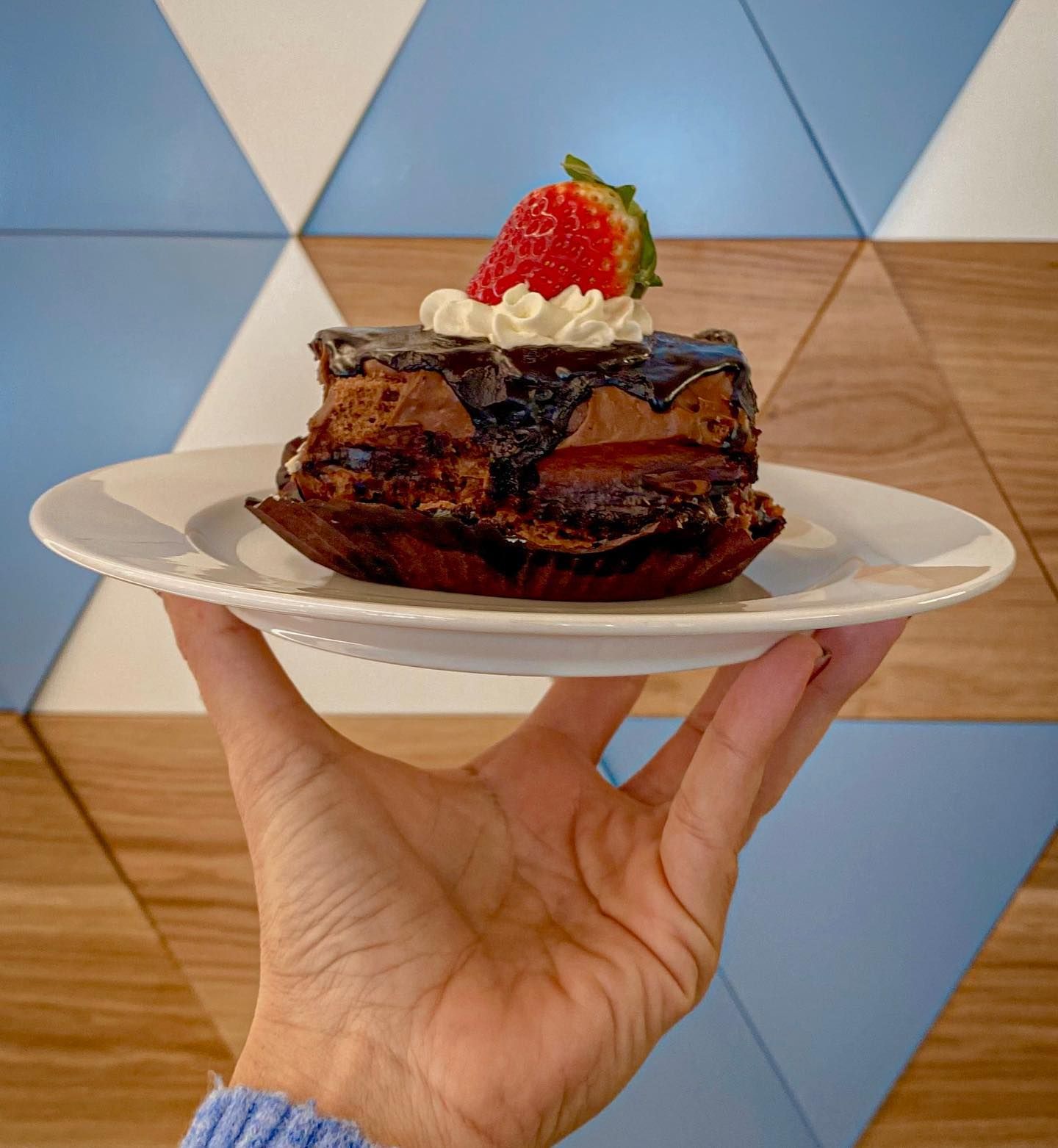 Mano che regge un piatto con una torta quadrata al cioccolato, guarnita con una fragola e panna montata.