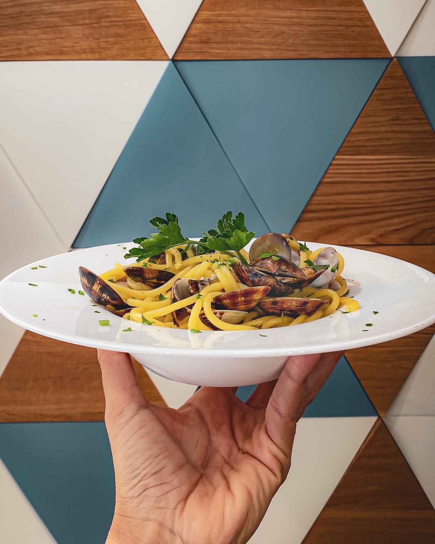 Mano che regge un piatto bianco di pasta con vongole e prezzemolo, contro una parete rivestita in legno con motivi geometrici.