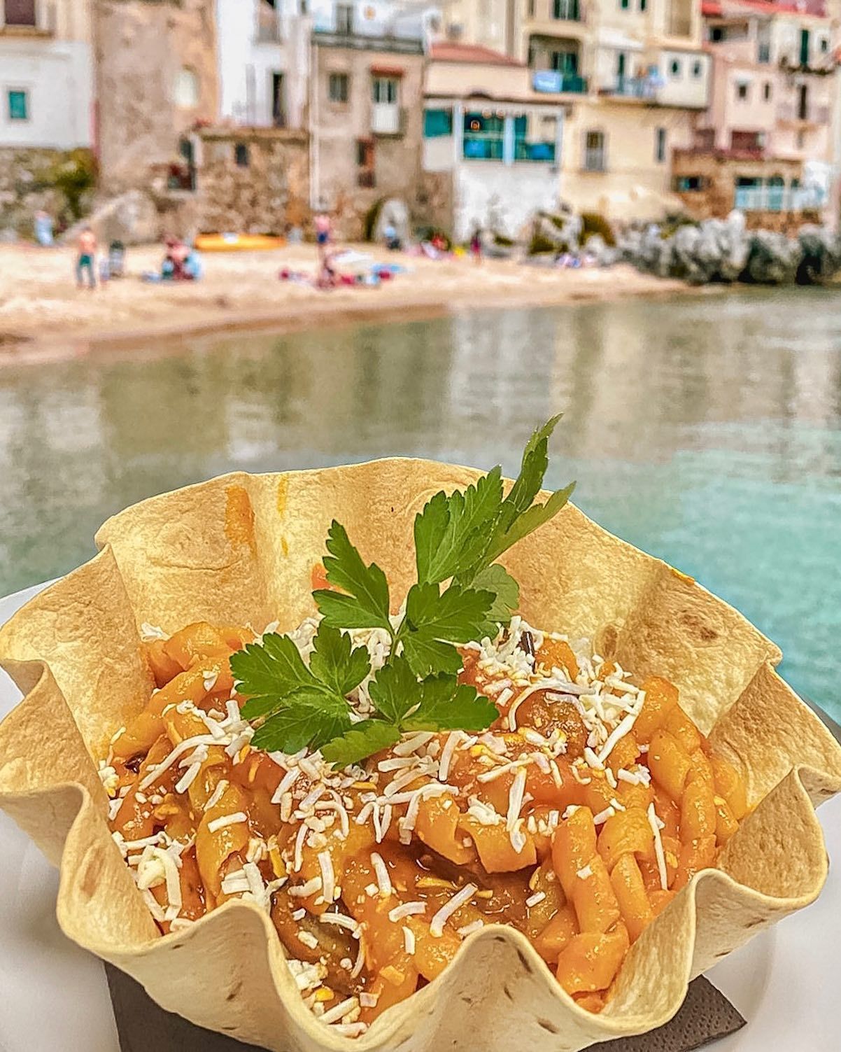 Piatto di pasta in una ciotola di tortilla, condito con formaggio e prezzemolo, spiaggia ed edifici sullo sfondo.