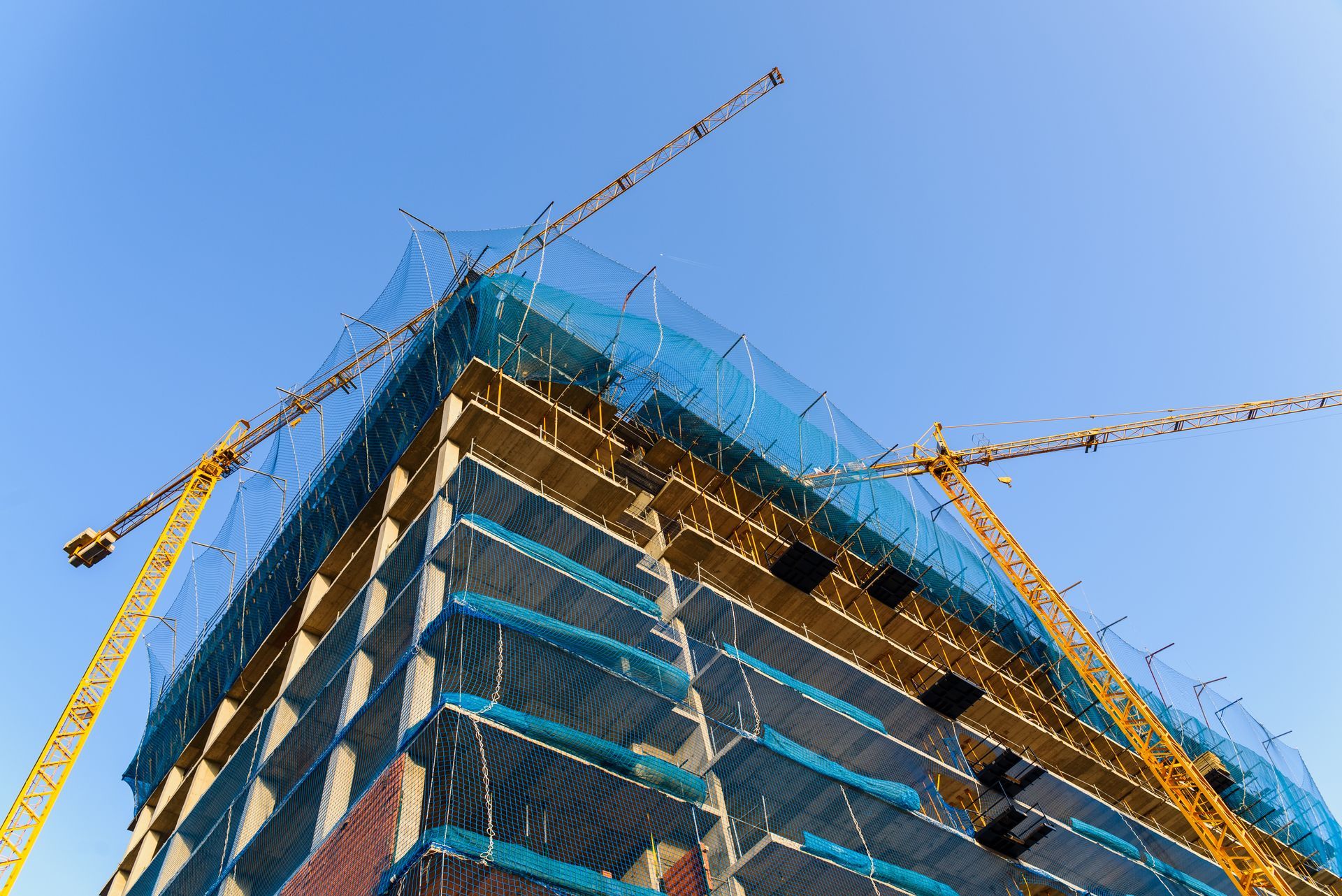 High-rise building under construction, wrapped in blue safety netting with cranes against the sky