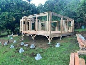 A wooden frame for a house under construction, resting on concrete piers in a grassy, wooded area.