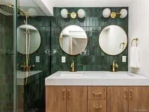 Modern bathroom vanity with two circular mirrors, green tile backsplash, wood cabinets, and gold fixtures.