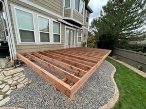 A partially constructed wooden deck frame attached to the back of a house above a gravel landscaped area.