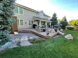 A tan two-story house with a wooden deck, stone patio, and grassy backyard under a clear blue sky.