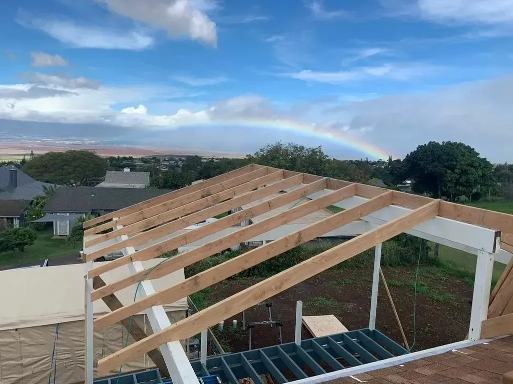 Wooden roof rafters under construction with a rainbow stretching across the sky above a neighborhood.