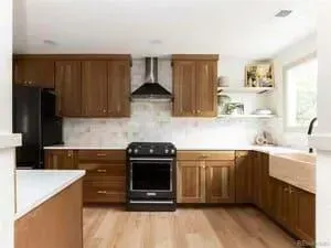 A modern kitchen featuring wooden cabinets, a black stove with a vent hood, white backsplash, and light wood flooring.