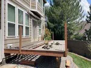 A partially built wooden deck attached to the side of a tan house with large windows and a gravel base below.