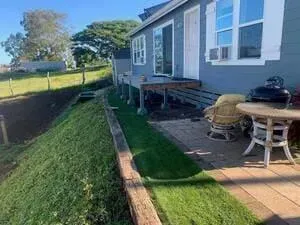 A blue house exterior with a wooden deck, a stone patio, and outdoor furniture next to a grassy slope.