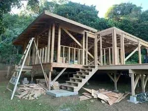 A wooden frame of a house under construction on stilts, featuring a staircase and surrounding trees.