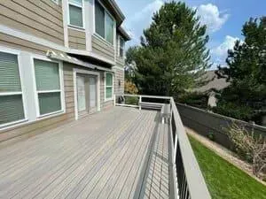 A wooden deck attached to the side of a beige two-story house with a white railing overlooking a lawn and trees.