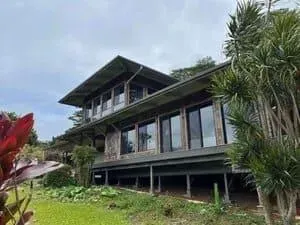 A modern two-story home with a dark exterior, wide roof overhangs, and large windows, surrounded by lush green foliage.