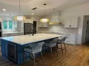 A bright kitchen featuring a large, blue-based island with white countertops, three gray stools, and pendant lighting.