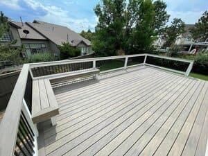 A spacious, grey-toned wooden deck with a built-in bench, white railings, and a view of neighboring houses and trees.