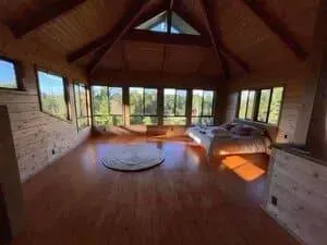 A spacious, sunlit wooden bedroom with high vaulted ceilings, a circular rug, and a bed facing large windows.