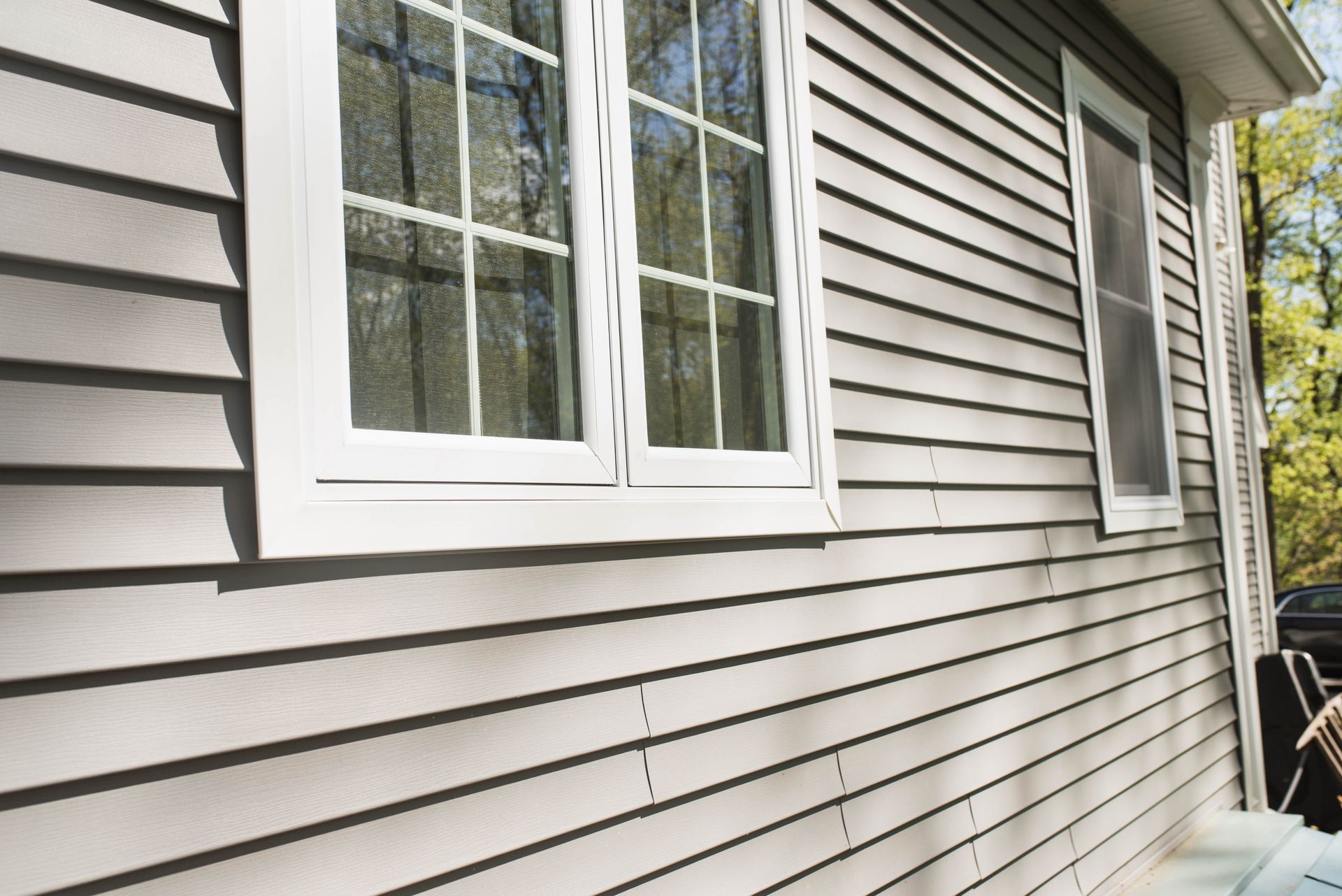 A close-up view of the side of a house with light gray vinyl siding, two white-framed windows, and white corner trim.