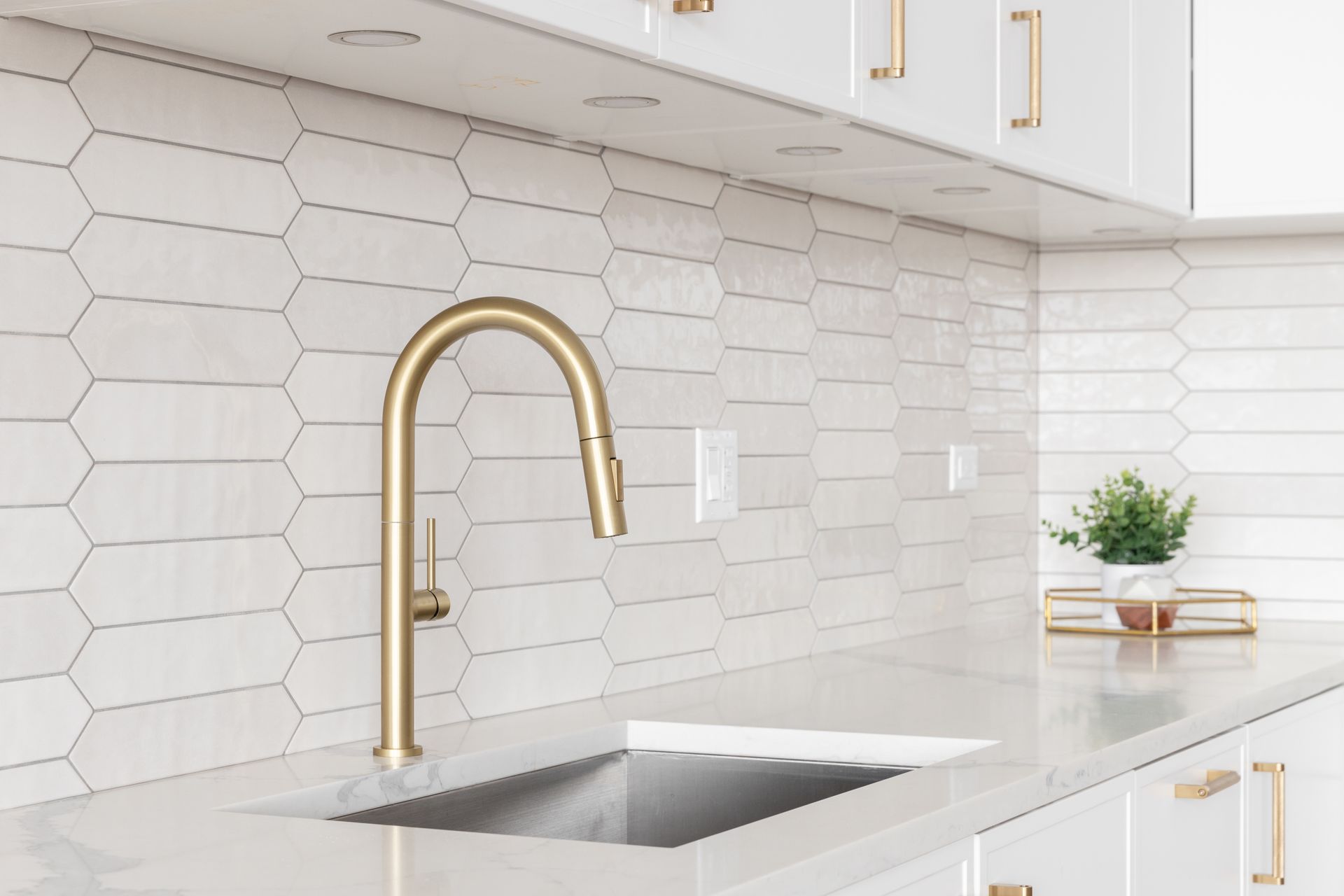 A gold faucet stands above a white kitchen sink against a light-colored, elongated hexagonal tile backsplash.