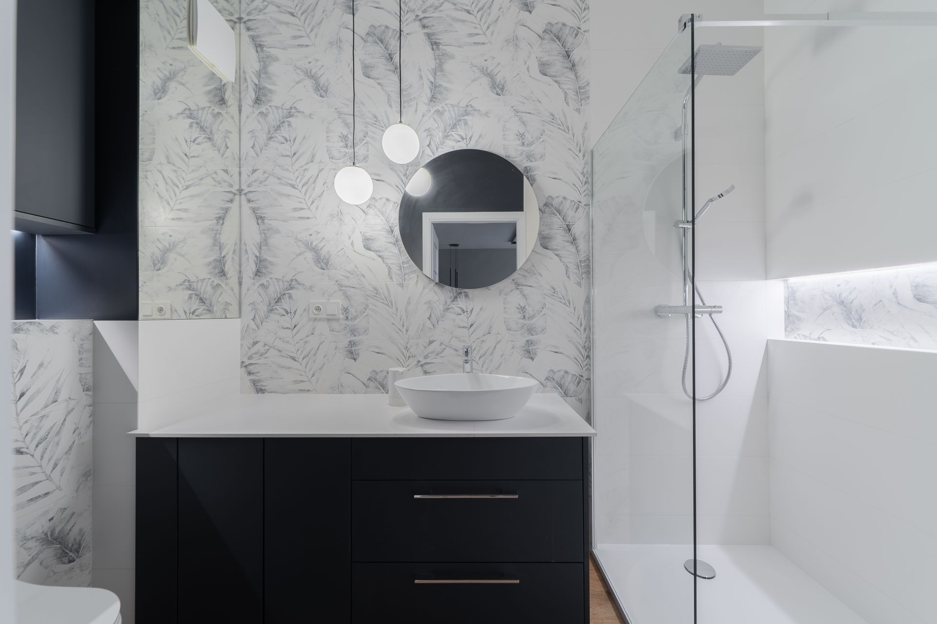 Modern bathroom with black vanity, white vessel sink, leaf-patterned tiled wall, round mirror, and glass walk-in shower.