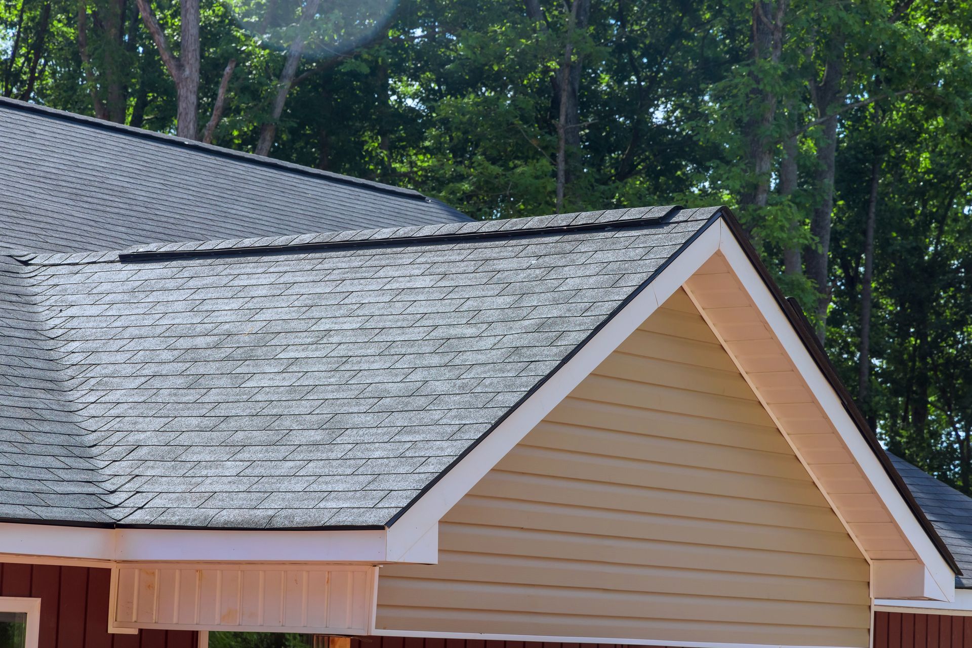 The gable and roofline of a house with gray shingles, tan siding, and white trim against a background of green trees.